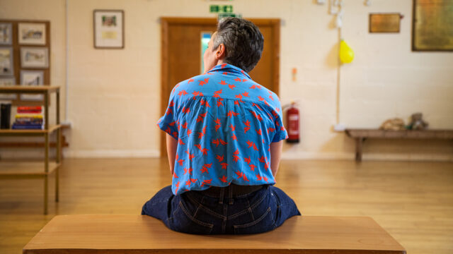 A white woman with short dark, greying hair, wearing a blue shirt with red flowers sirs on a table with her back facing the camera