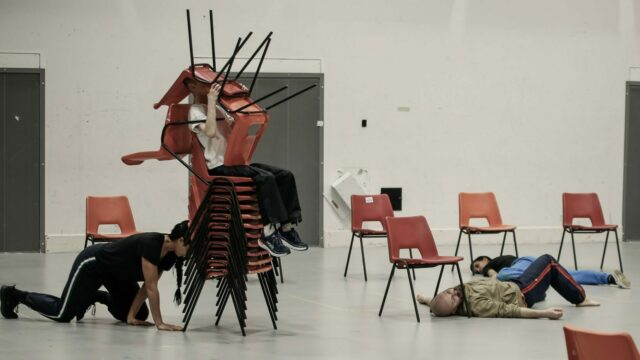 A person sits on a tall stack of chairs with two more chair upside down, covering their face. A mixed race woman with long dark braids pushes the stack of chairs on all fours with their head. There are empty chairs scattered around the room. There are two more people sprawled across the floor, one on their front and one on their back.