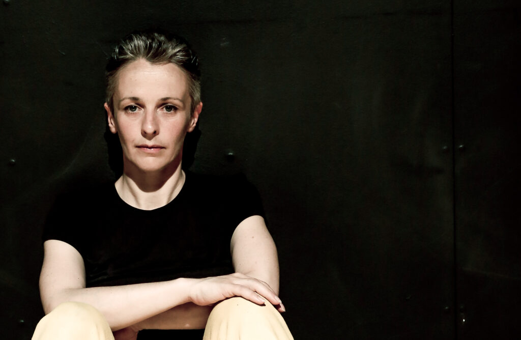 A woman with short dark, greying hair sits on the floor with her feet on the ground with her arms across her knees. The room is black, she wears a black T shirt and looks calmly straight into the camers