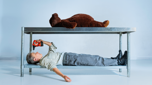 A large teddy bear lies onto of a stainless steel table. A white woman, in grey workwear lies on the shelf under the table. She is holding a toy drill to her head.